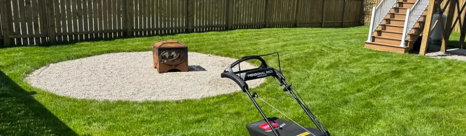 Mown lawn with a round gravel pad and fire pit, a wooden fence behind and a red push mower in front